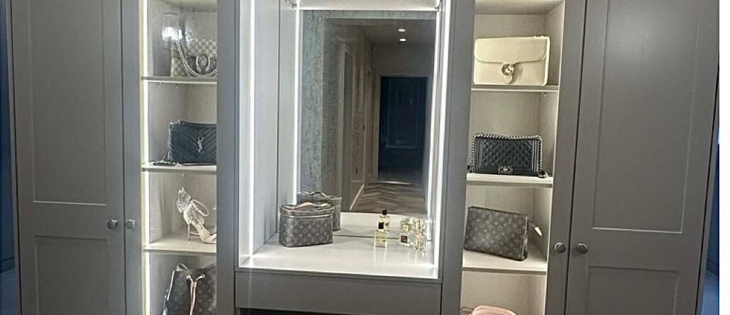 Custom-built wardrobe with integrated lighting, vanity area, and shelving units for storage of bags, shoes, and accessories in a contemporary dressing room. Built in Wardrobes Suffolk